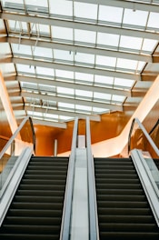An upward view of an escalator with modern design and sleek lines. The ceiling features large windows, allowing natural light to illuminate the space. The walls have a warm, amber tone reflecting off the surfaces.