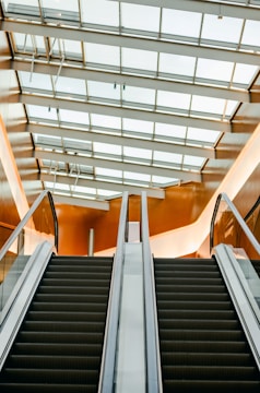 An upward view of an escalator with modern design and sleek lines. The ceiling features large windows, allowing natural light to illuminate the space. The walls have a warm, amber tone reflecting off the surfaces.