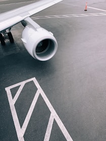 A close-up view of an airplane's wing and engine on the tarmac, accompanied by ground markings and a single orange traffic cone in the background.