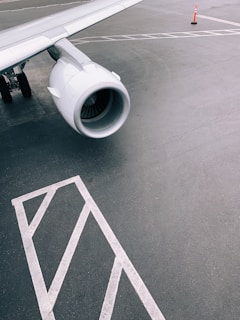 A close-up view of an airplane's wing and engine on the tarmac, accompanied by ground markings and a single orange traffic cone in the background.