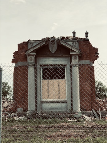 An old, partially demolished brick building with classical architectural details is situated behind a chain-link fence. The facade includes two columns and a pediment, and the top appears to have been removed or damaged. Debris surrounds the structure, indicating an ongoing demolition process.