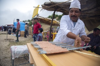 A man wearing a white chef's uniform and hat is preparing food on metal skewers. He is part of an outdoor setting with several people in the background, some of whom are standing near makeshift stalls and tents. The environment appears to be a market or festival with a rustic and informal atmosphere.