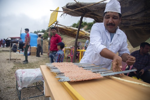 A man wearing a white chef's uniform and hat is preparing food on metal skewers. He is part of an outdoor setting with several people in the background, some of whom are standing near makeshift stalls and tents. The environment appears to be a market or festival with a rustic and informal atmosphere.