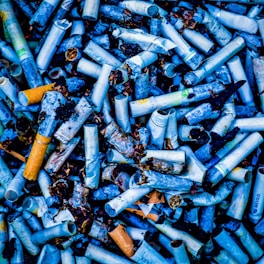 A densely packed collection of discarded cigarette butts, some stained and partially burnt, scattered randomly. The image features various brand markings and colors on the cigarette filters, set against a backdrop of dark and stained tobacco remnants.
