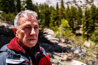 An older man with short gray hair is standing outdoors, wearing a red and black jacket. He appears contemplative and is surrounded by a forested area with evergreen trees in the background. Sunlight casts shadows on his face, indicating a clear, bright day.