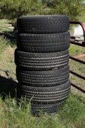 A stack of six black tires stands vertically on grassy ground. The background features a mixture of green foliage and some metal structures. The tires display visible treads and signs of wear.