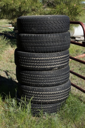 A stack of six black tires stands vertically on grassy ground. The background features a mixture of green foliage and some metal structures. The tires display visible treads and signs of wear.