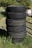 A stack of six black tires stands vertically on grassy ground. The background features a mixture of green foliage and some metal structures. The tires display visible treads and signs of wear.