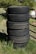 A stack of six black tires stands vertically on grassy ground. The background features a mixture of green foliage and some metal structures. The tires display visible treads and signs of wear.