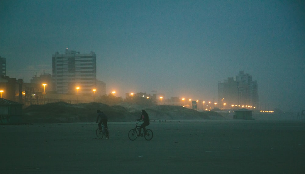 two people biking on snow field viewing high-rise buildings during night time