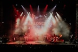 A live music performance with a band on stage surrounded by red and orange stage lights. Smoke fills the air, creating a dramatic atmosphere as the band members play their instruments under a colorful backdrop.