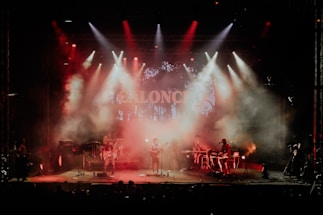 A live music performance with a band on stage surrounded by red and orange stage lights. Smoke fills the air, creating a dramatic atmosphere as the band members play their instruments under a colorful backdrop.