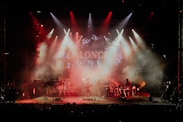 A live music performance with a band on stage surrounded by red and orange stage lights. Smoke fills the air, creating a dramatic atmosphere as the band members play their instruments under a colorful backdrop.