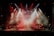 A live music performance with a band on stage surrounded by red and orange stage lights. Smoke fills the air, creating a dramatic atmosphere as the band members play their instruments under a colorful backdrop.