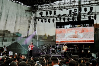 A large outdoor concert stage featuring a band performing in front of a crowd. The stage setup includes multiple musical instruments such as drums and guitars. A backdrop screen displays an image of a brick wall with text on it. Spotlights and stage lights are visible overhead.