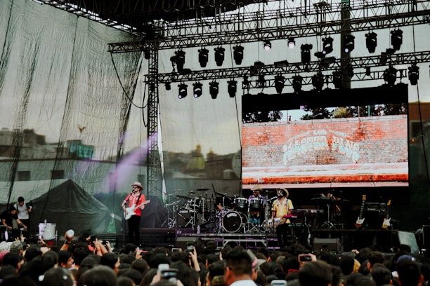 A large outdoor concert stage featuring a band performing in front of a crowd. The stage setup includes multiple musical instruments such as drums and guitars. A backdrop screen displays an image of a brick wall with text on it. Spotlights and stage lights are visible overhead.