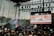 A large outdoor concert stage featuring a band performing in front of a crowd. The stage setup includes multiple musical instruments such as drums and guitars. A backdrop screen displays an image of a brick wall with text on it. Spotlights and stage lights are visible overhead.