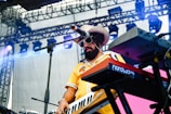 A musician with a beard and sunglasses plays a keyboard on stage. They are wearing a yellow shirt and a large hat. The setting includes stage lighting and equipment visible in the background.