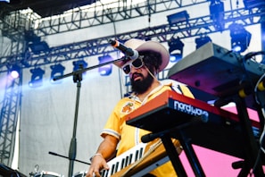 A musician with a beard and sunglasses plays a keyboard on stage. They are wearing a yellow shirt and a large hat. The setting includes stage lighting and equipment visible in the background.
