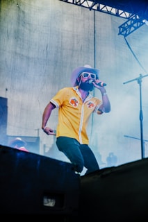 A musician wearing a bright yellow shirt, white cowboy hat, and sunglasses performs on stage under a metal frame structure. The scene is filled with smoke, suggesting the use of stage effects, and the performer holds a microphone close to his mouth.