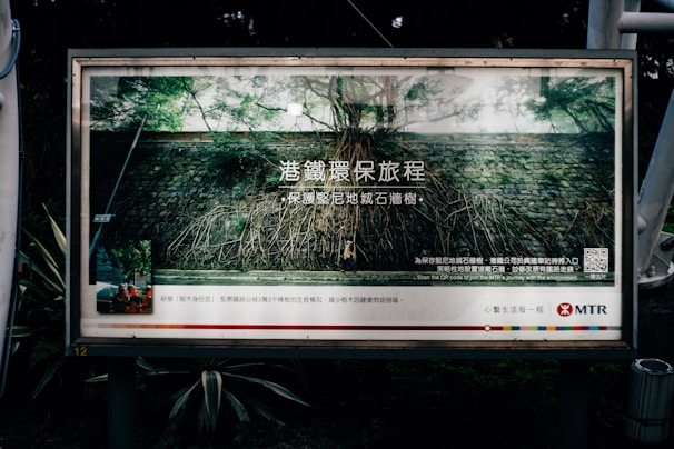 A public advertisement poster situated outdoors, featuring a stone wall with tree roots visible on the surface. The poster includes text in Chinese and English, along with a QR code and the MTR logo. The surrounding environment includes vegetation and metal structures.