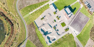 An aerial view of a skate park with various ramps and obstacles, next to a basketball court. Surrounding the park are well-maintained grassy areas and a parking lot with cars. A curving path and patches of vegetation are visible on the left side.