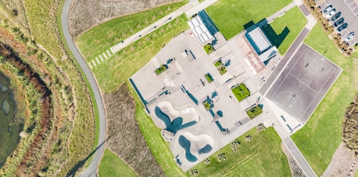 An aerial view of a skate park with various ramps and obstacles, next to a basketball court. Surrounding the park are well-maintained grassy areas and a parking lot with cars. A curving path and patches of vegetation are visible on the left side.
