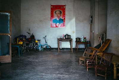 A simple room with a concrete floor and gray walls, featuring a poster of a notable figure above a small wooden table. The room contains a few chairs, a table with a kettle and other household items. Bicycles and a motorbike are parked along one wall, and a bamboo recliner is placed near some chairs.