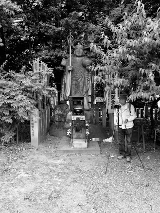 A large stone statue stands amid lush foliage, holding a staff and adorned with traditional garments. A man is positioned to the right, adjusting a tripod, indicating a photography setting. Several signs and banners with Japanese characters flank the statue, suggesting a cultural or religious site. The image is in black and white, enhancing the contrast between the natural and man-made elements.