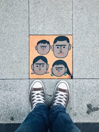 A pair of sneakers on a tiled pavement, with a colorful tile featuring abstract faces of four people, two adults and two children, arranged in a family-like manner.