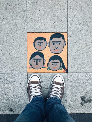 A pair of sneakers on a tiled pavement, with a colorful tile featuring abstract faces of four people, two adults and two children, arranged in a family-like manner.