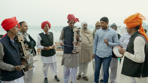 A group of men, some wearing traditional attire with red and orange turbans, are gathered together playing musical instruments. One man appears to be singing while others play string instruments. The background shows a waterfront or riverside setting, suggesting an outdoor gathering.