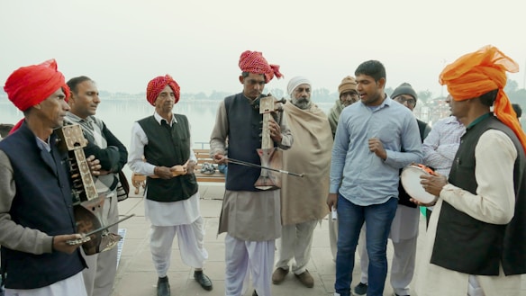 A group of men, some wearing traditional attire with red and orange turbans, are gathered together playing musical instruments. One man appears to be singing while others play string instruments. The background shows a waterfront or riverside setting, suggesting an outdoor gathering.