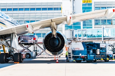 A large commercial airplane is parked on an airport tarmac with a jet bridge connected to it. The aircraft is being serviced with fuel trucks and other ground support equipment nearby. The airplane's wing and engine dominate the foreground, and there are several maintenance workers around the ground vehicles.