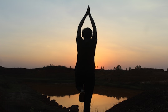 A silhouette of a person standing on one leg in a yoga pose against a sunset backdrop. The sky is a gradient of soft oranges and blues, and the horizon features a calm landscape with trees and a body of water reflecting the colors of the sky.