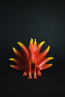 A stylized fox-like figurine with multiple tails is centered against a dark background. The figure is primarily red with bright orange and yellow highlights on the tips of its tails and ears, creating a vibrant contrast.