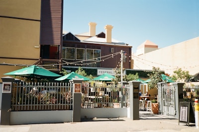 An outdoor café or restaurant area with multiple tables under green umbrellas with Heineken logos. The space is surrounded by a metal fence, and string lights are hung across above the seating area. There is a multi-story building with a mix of wooden and plaster walls in the background, with visible chimneys and windows. Some plants and small trees are also present within the fenced area.