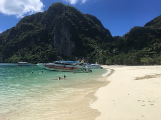 Several speedboats are docked near a sandy beach with clear turquoise water. People are swimming in the water while lush green hills rise in the background under a clear blue sky.
