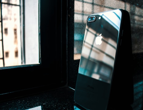 A smartphone with a reflective back displaying the Apple logo sits on a dark, glossy surface near a window. The window frames and some of the outdoor architectural elements are visible through the reflection.