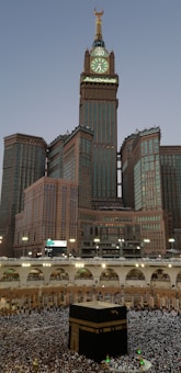 A large, ornate clock tower with a crescent moon on top dominates the skyline, surrounded by skyscrapers. Below, a massive crowd encircles a cubic, black structure known as the Kaaba in an open courtyard.