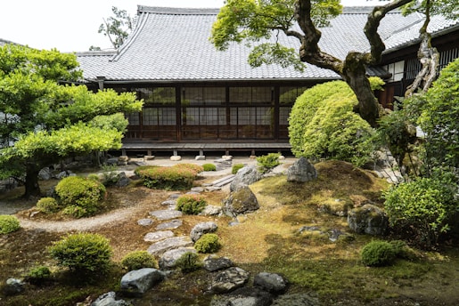 A tranquil Japanese garden with neatly arranged bushes, stones, and a meandering stone path. A traditional wooden building with large windows forms the backdrop, and lush greenery with a variety of plants is abundant. The garden appears meticulously cared for, embodying a serene and harmonious atmosphere.