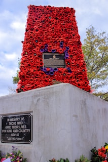 A war memorial monument, elaborately decorated with vibrant red poppies and some purple accents. Two plaques are visible on the monument, with inscriptions commemorating those who gave their lives for their country. The base is made of concrete, with additional floral tributes laid at the bottom. Surrounding the monument are green trees and a blue sky above.