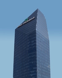 A modern skyscraper with a glass facade and the logo of BNP Paribas displayed prominently at the top against a clear blue sky. The building is characterized by its rectangular shape with vertical lines, creating a sleek and professional appearance.