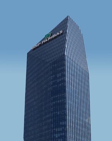 A modern skyscraper with a glass facade and the logo of BNP Paribas displayed prominently at the top against a clear blue sky. The building is characterized by its rectangular shape with vertical lines, creating a sleek and professional appearance.