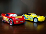Two colorful toy cars are placed on a wooden surface. The car on the left is red with number 95 and features bold graphics on its sides, including bright yellow flames. The car on the right is yellow with blue accents, displaying the number 51 and the word 'Dinoco' prominently. The cars have eyes on their windshields, giving them a cartoonish, character-like appearance.