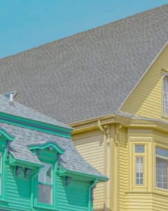 The image features two adjacent houses with distinct and vibrant colors. The house on the left is painted a bright teal color with intricate trim details and a gray shingled roof, while the house on the right is a pastel yellow with a matching roof. Both houses exhibit architectural charm, suggesting a residential neighborhood.