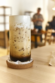 A tall glass filled with iced coffee or bubble tea sits on a wooden table, with a frothy top and dark layers at the bottom, placed on a cork coaster with a lace doily. The background is a soft-focus cafe interior where a person is seen standing, adding a casual and relaxed atmosphere.