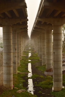 A symmetrical arrangement of large concrete pillars supporting an elevated structure, possibly a bridge or a highway. There is a canal or drainage system running between the rows of pillars, with patches of green vegetation along its edges. The scene is enveloped in a hazy atmosphere.