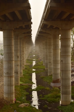 A symmetrical arrangement of large concrete pillars supporting an elevated structure, possibly a bridge or a highway. There is a canal or drainage system running between the rows of pillars, with patches of green vegetation along its edges. The scene is enveloped in a hazy atmosphere.