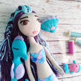 A handmade fabric mermaid doll with long, multicolored yarn hair and a blue and pink scaled top. The doll is lying on a textured wooden surface along with a blue seahorse plush, a small fish plush, and spools of sewing thread in shades of blue and pink. The doll has large eyes with painted details and is accompanied by a seashell accent.